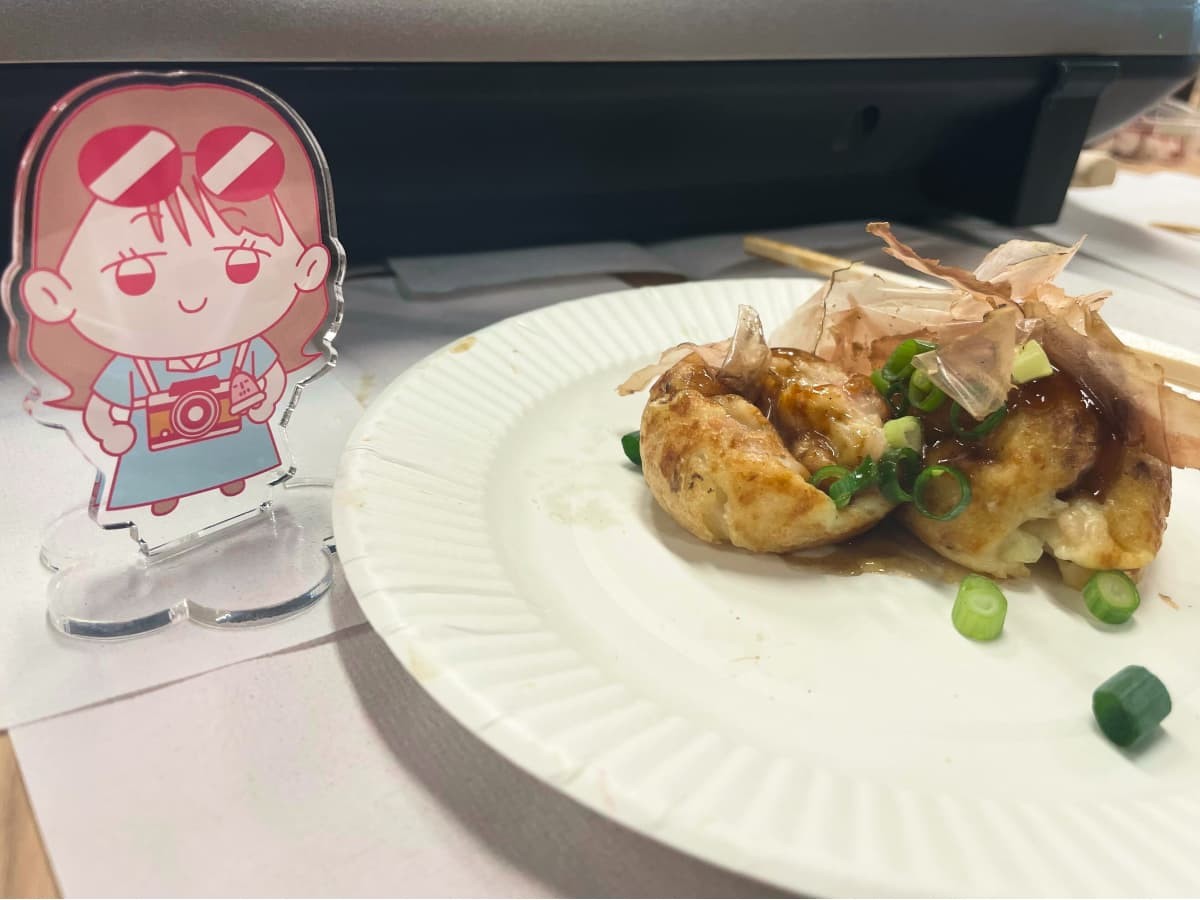 女性社員「ヨネチ」のイラストアクリルスタンドの横に、紙皿に乗ったたこ焼きが置かれている。ヨネチは頭にサングラスをかけ、水色のワンピースを着て首からカメラを下げ微笑んでいる。