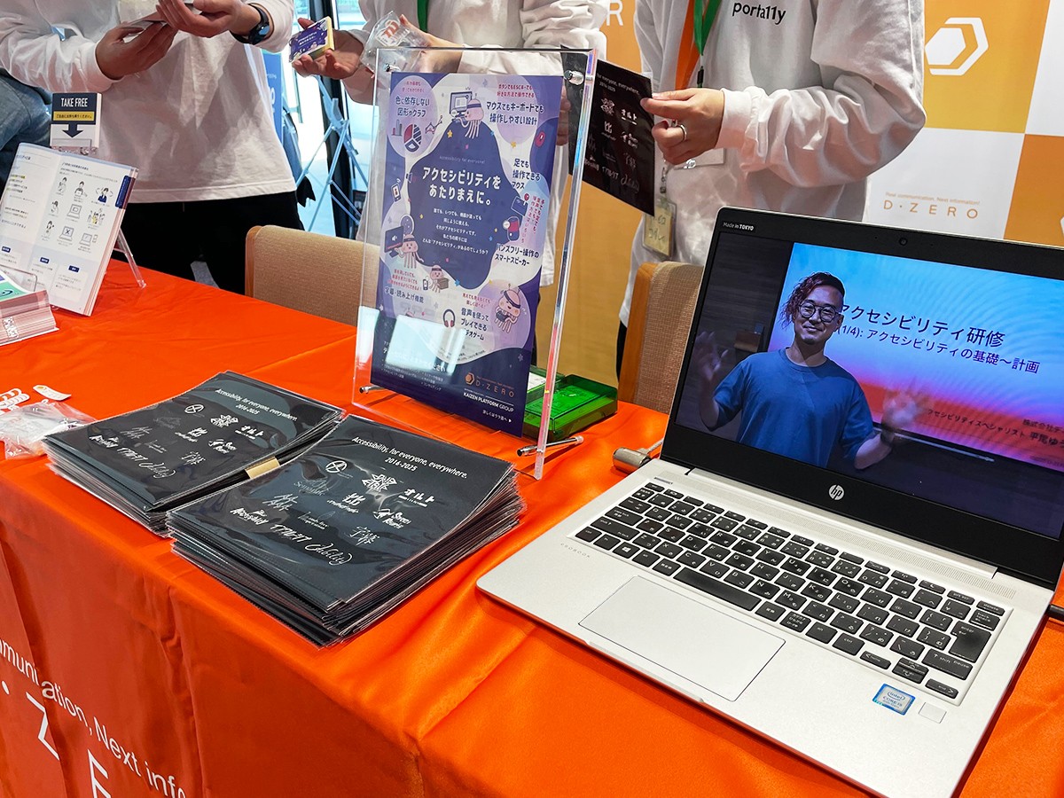 ディーゼロのスポンサーブースの机の様子。オレンジ色のテーブルクロスが敷かれた机上に、ディーゼロのCM動画を再生中のノートパソコンやノベルティのマイクロファイバークロスなどが置かれている。