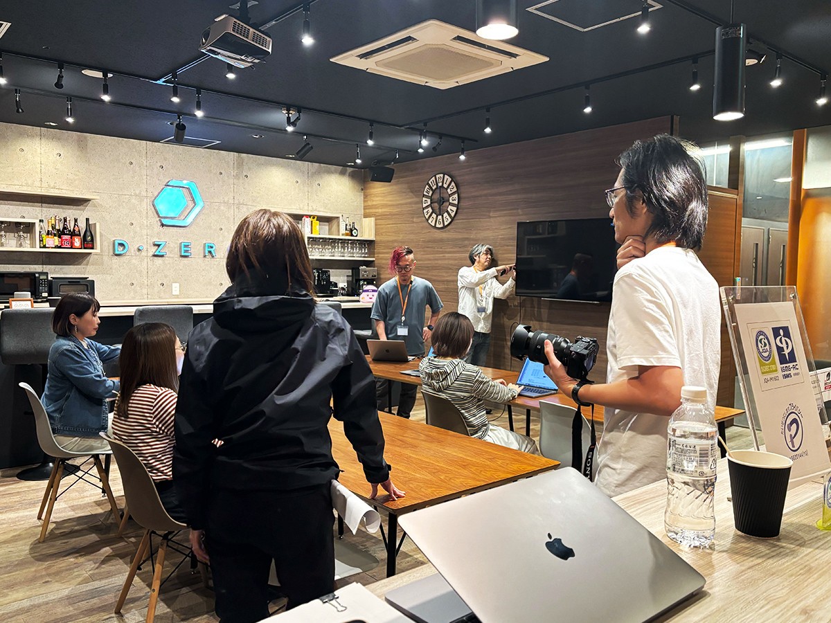 ディーゼロオフィスのカフェスペースでCM動画の撮影メンバーが集まり、モニターやPC、カメラなどの撮影準備をしている。