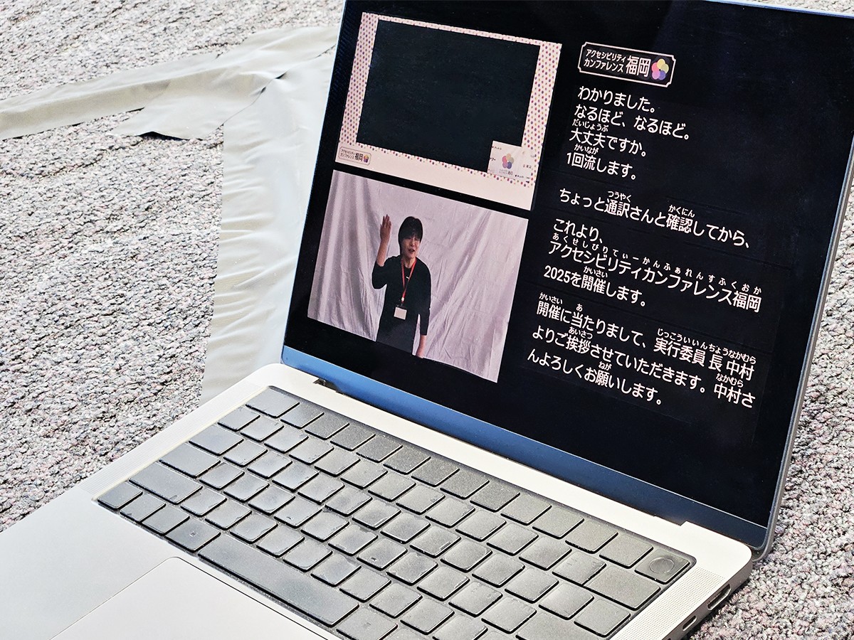 ノートパソコンに表示された情報保障版のYouTube配信画面。画面右半分にはリアルタイム字幕が大きく表示され、左側には上部に会場の配信、下部に手話通訳者の映像が表示されている。