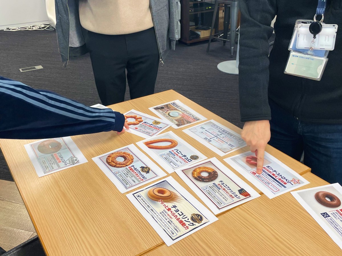 各ドーナツの特徴が記載された紙がテーブルの上に並べられており、スタッフ2名が食べたいドーナツの紙に指を指している。