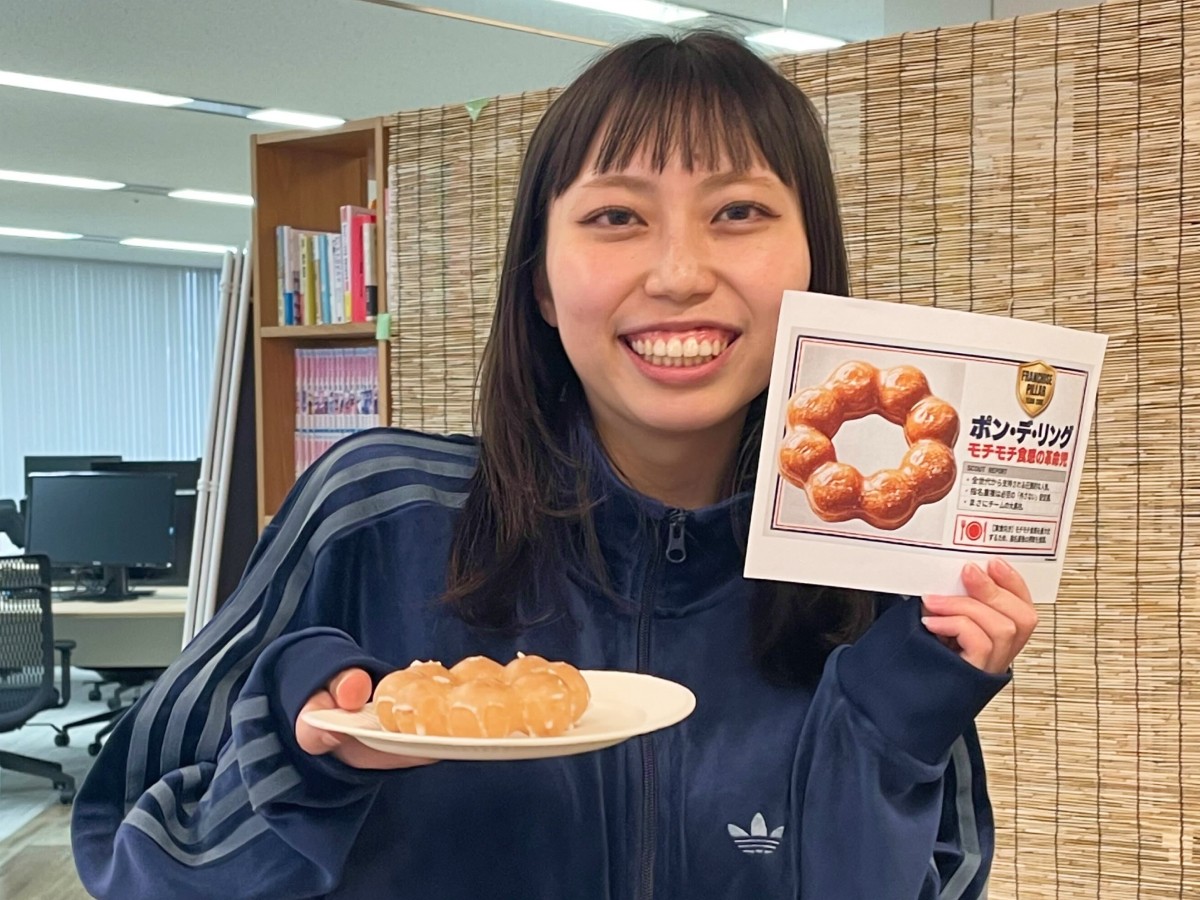 女性スタッフが右手にドーナツ、左手にそのドーナツの詳細が書かれた紙を持って、笑顔でカメラの方を向いている。